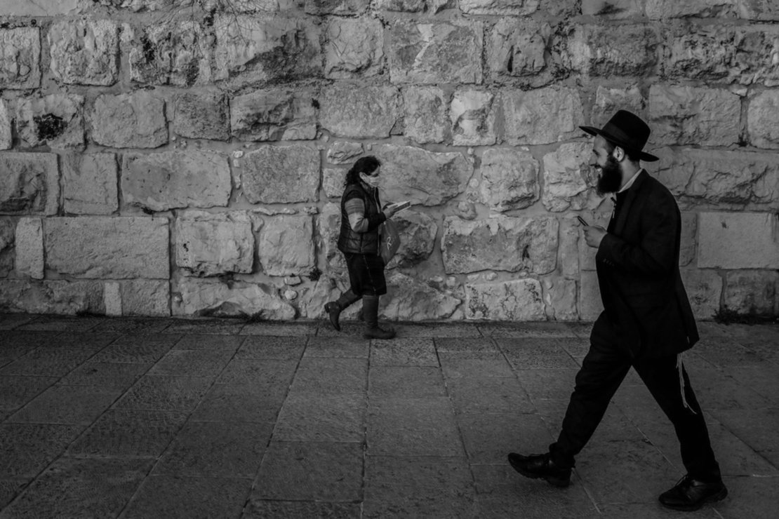 Una mujer con una máscara facial protectora y un hombre judío ortodoxo pasan cerca de los muros de la Ciudad Vieja en Jerusalén. Una mujer con una máscara facial protectora y un hombre judío ortodoxo pasan cerca de los muros de la Ciudad Vieja en Jerusalén.